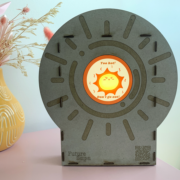 A laser cut UV indicator clock made of cardboard sits atop a pink cabinet next to a yellow vase holding pink and white dried flowers. The UV clock indicates it is too hot to go outside with an animation of an angry hot sun.
